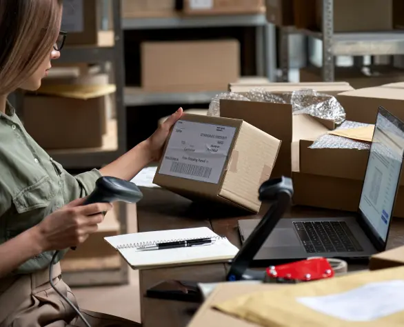 Mulher segurando um leitor de código de barras em uma mão e uma caixa de papelão na outra em uma mesa à frente de um notebook