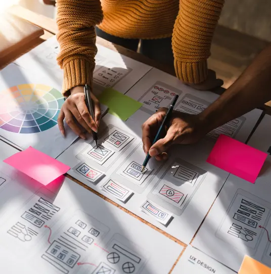 Mulher de pé com uma caneta em sua mão desenhando uma interface gráfica em um papel sobre a mesa