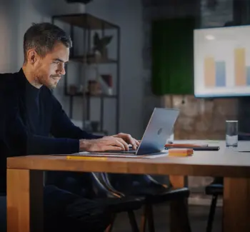 Um homem sentado em uma mesa em frente à um notebook