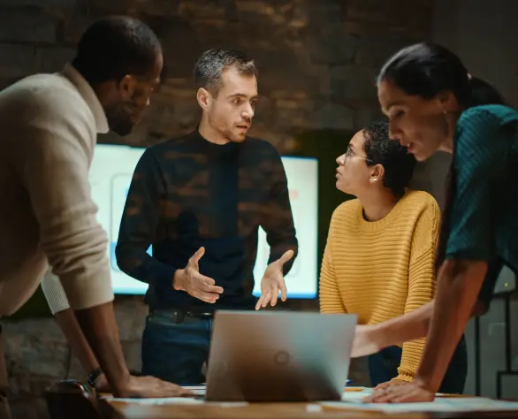 Quatro pessoas em pé conversando reunidas ao redor de uma mesa com um notebook