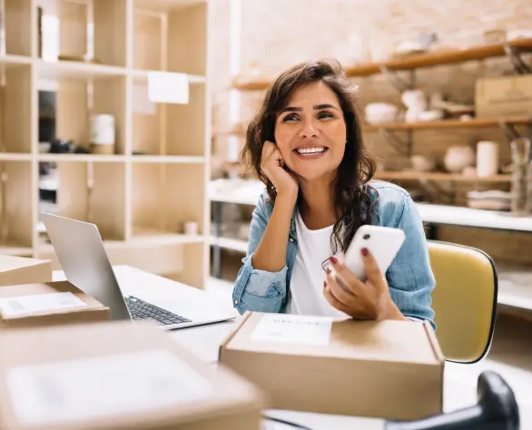 Mulher sorrindo sentada em uma mesa com um notebook, caixas e segurando um celular